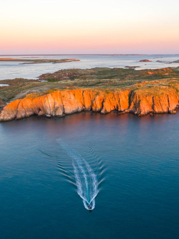 A boat travels on the serene, calm waters of the Kimberley sea towards Cygnet Bay Pearl Farm. In the background is an island, part of the Archipelago of islands, glowing in orange hues from the stunning sunset. The sunset lights up the sky in a warm gradient, in contrast with the azure waters in the foreground.