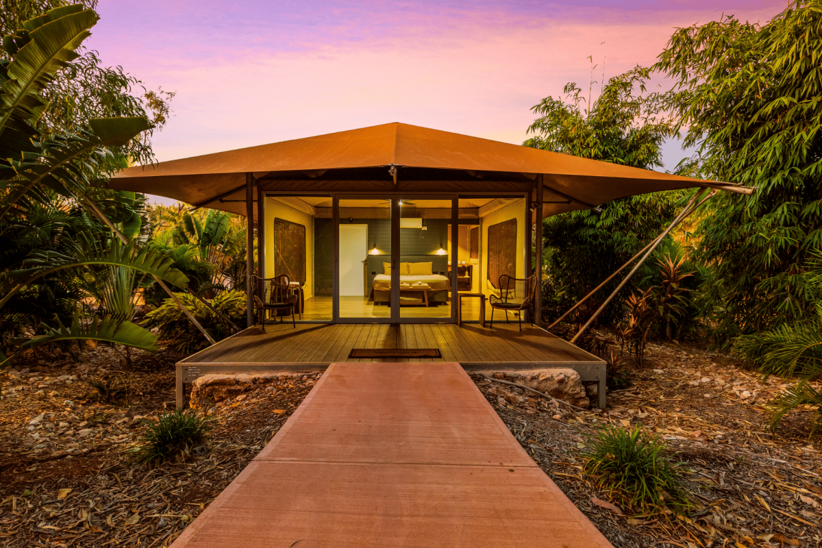 A path leads to a Pearler's Village Safari Tent glowing from within at dusk. At this time of the day you can see inside the tent, showing the comfortable king bed with two ambient lamps on either side of the bed, the door to the ensuite of the tent and the passage to the self countained kitchenette area as well. Behind the tent is a beautiful Kimberley sunset, with pink and purple hues, and surrounding the tent are native, tropical plants - creating serenity and calm for those staying in this accommodation.