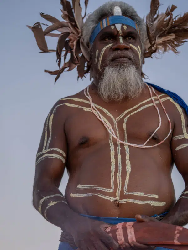 A Bardi Jawi man is dressed in traditional clothing, with culturally significant body paint covering his face, chest and arms. He wears a headband with a piece of Mother of Pearl resting between his forehead and the headband, shining with great lustre, and at the back of his head are several feathers that flourish from the sides of his head. He holds a boomerage in both hands and stares into the distance.