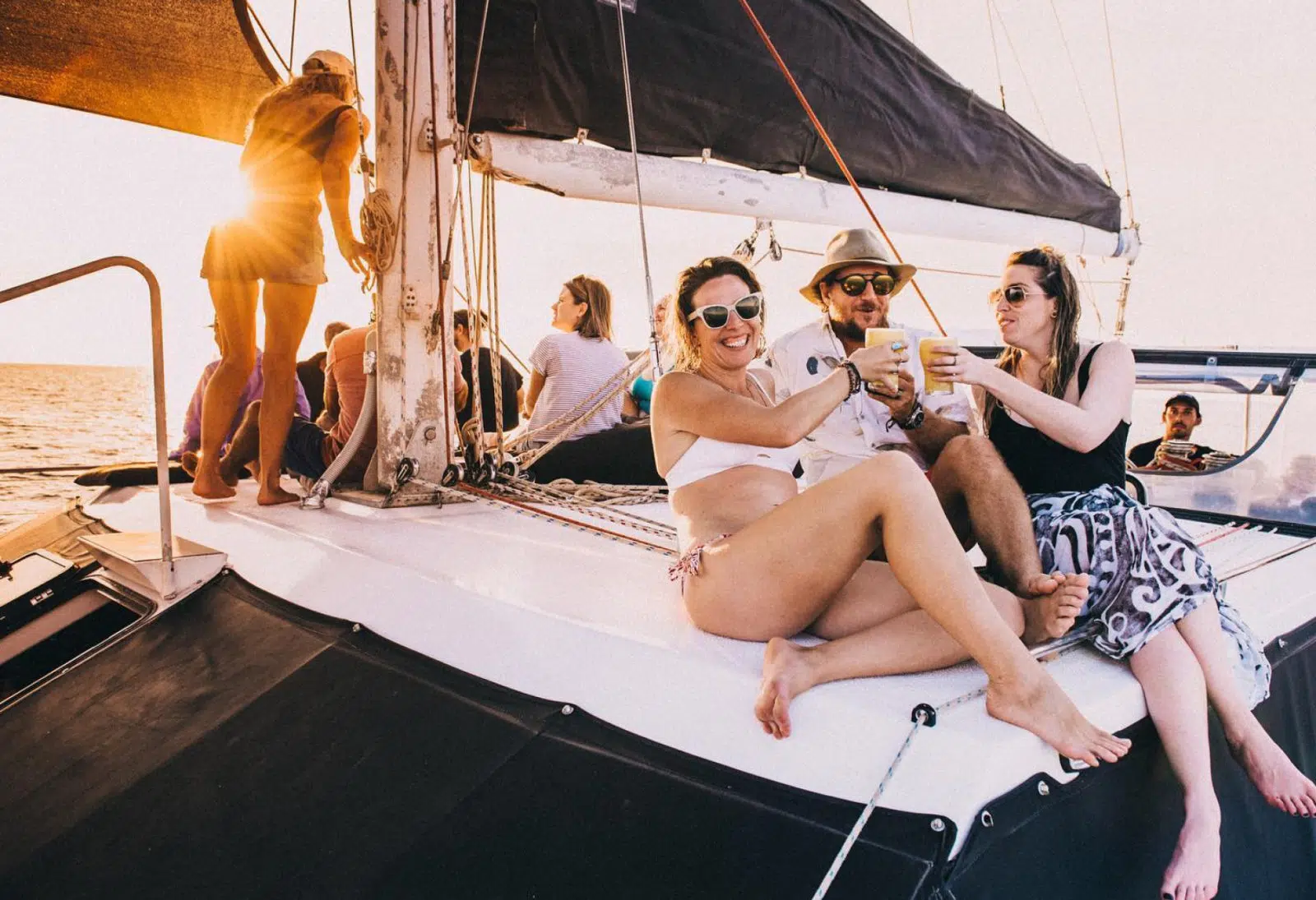 A group of guests sit on a boat, laughing and clinking their drinks. In the distance is the stunning sunset, and the blue waters known to Broome and the Kimberley.