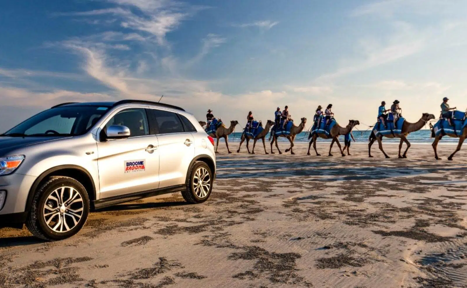 A 4WD sits on a white sandy Broome beach, with the waves crashing in the background and a line of camels with tourists sitting on them, enjoying Cable Beach.