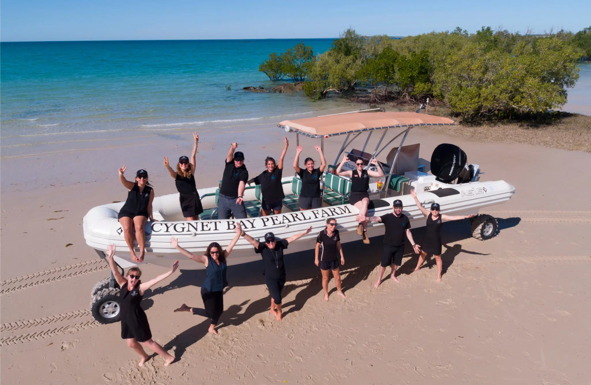 Our Cygnet Bay Pearl Farm team surround a boat used for tours, all posing and smiling while stand on the beach. In the background is the iconic blue waters of the Kimberley Coastline. Our Community.