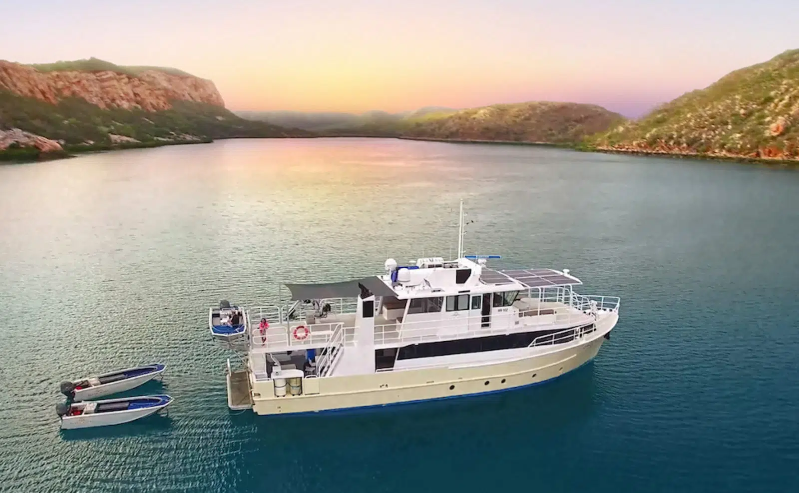 A cruise sits on the still Kimberley waters in the foreground, while guests watch a beautiful sunset in the background.