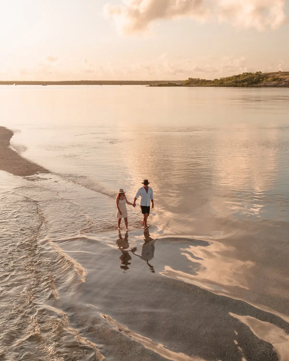 A couple walk hand in hand at sunset on the shoreline of a secret island. The sea is so still that it reflects that couple, the beautiful white clouds, and the peachy tones of the sunset. In the distance is the coastline of the Dampier Peninsula, with sandy white beaches. Beyond the Pearl Farm on The Dampier Peninsula.
