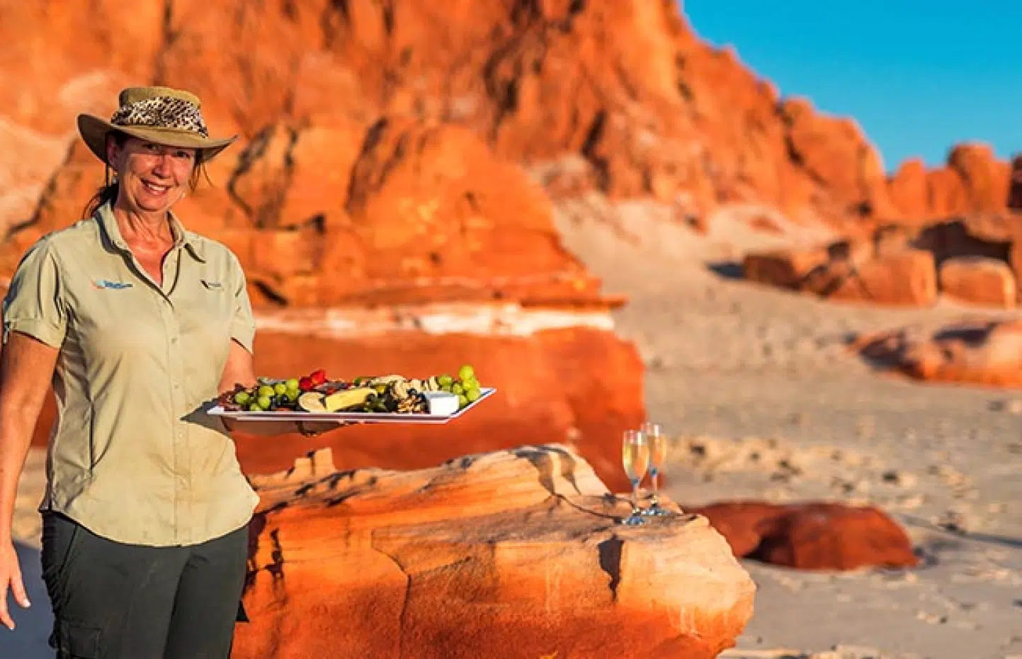 A tour guide holds a charcuterie board of grapes, cheeses and crackers on a stunning Kimberley beach, secluded and remote. In the distance are the iconic red rocks by the white sandy beach.