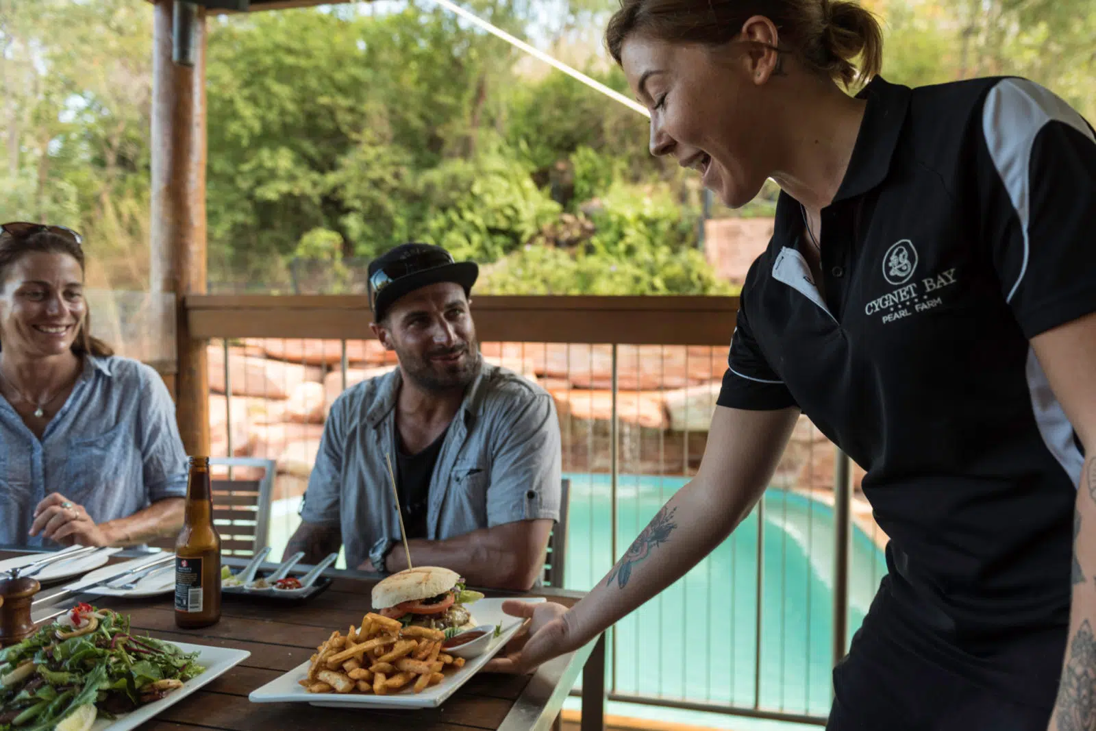 A service person puts down a burger and chips for guests attending lunch at the Homestead Restaurant during their stay at Cygnet Bay Pearl Farm. In the background is the infinity pool.