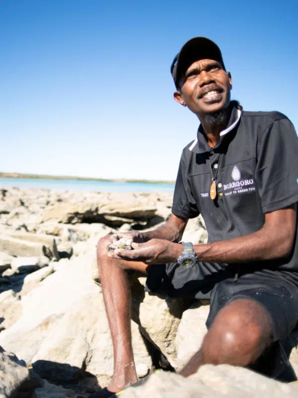 Terry Hunter sits on a beach during his Borrgoron Cultural Tour. Behind him is the shoreline of Cygnet Bay.