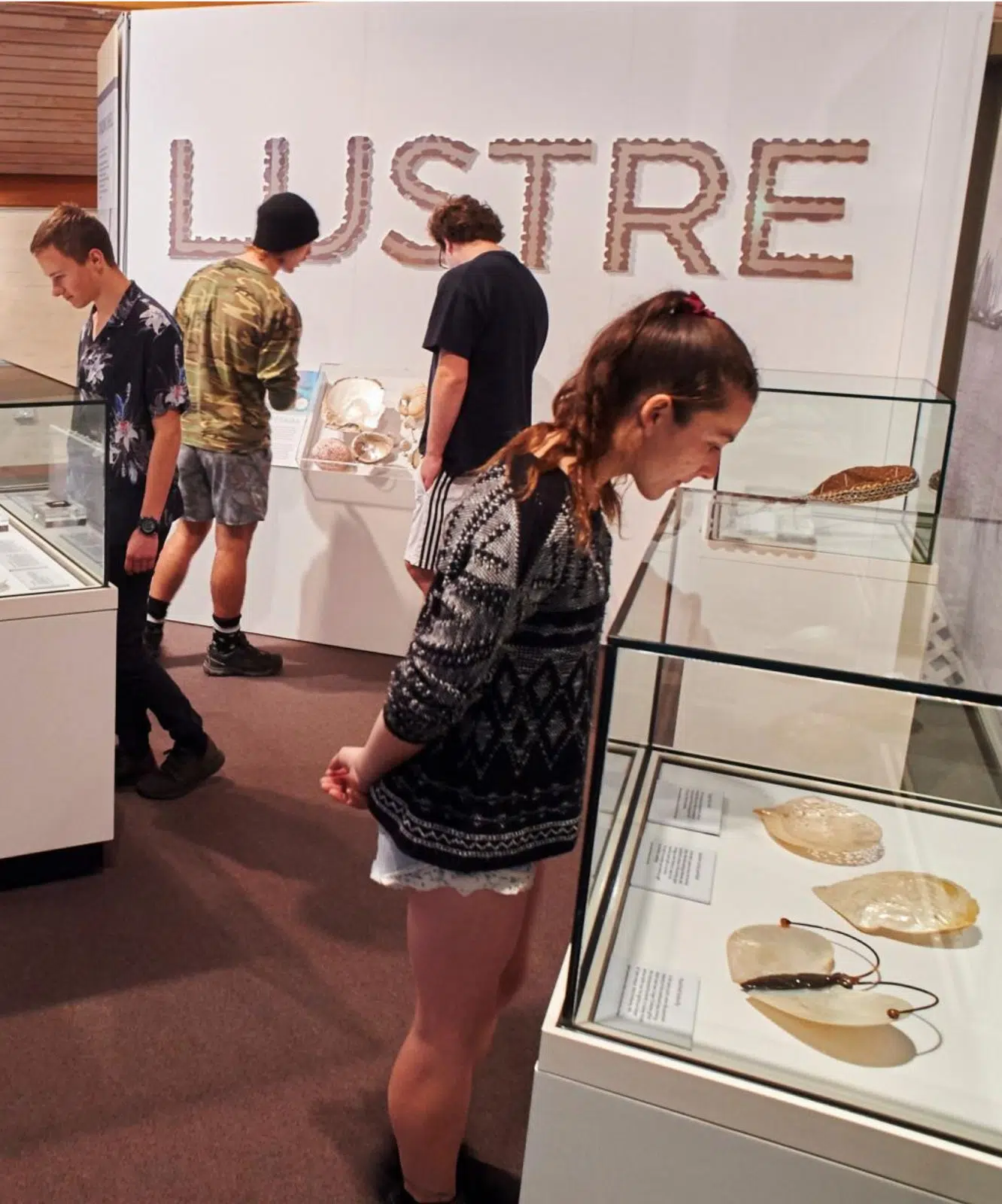 Individuals look into glass cabinets, reading about the pieces on display at the Lustre exhibition.