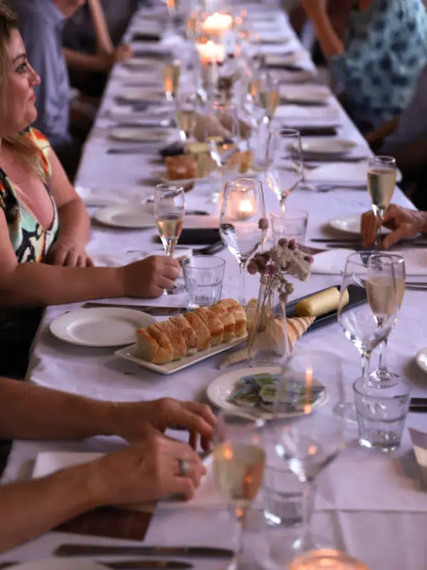 A group sit intimately at a table at sunset, overlooking the beautiful ocean of the Dampier Peninsula. The table is set with beautiful coastal elements of shells, and native florals, as well as candles to create a lovely, ambient setting.