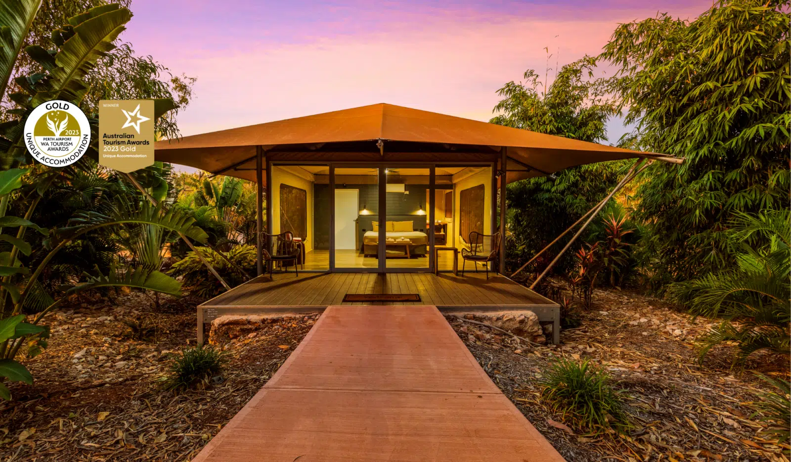 A path leads to a Pearler's Village Safari Tent glowing from within at dusk. At this time of the day you can see inside the tent, showing the comfortable king bed with two ambient lamps on either side of the bed, the door to the ensuite of the tent and the passage to the self countained kitchenette area as well. Behind the tent is a beautiful Kimberley sunset, with pink and purple hues, and surrounding the tent are native, tropical plants - creating serenity and calm for those staying in this accommodation.