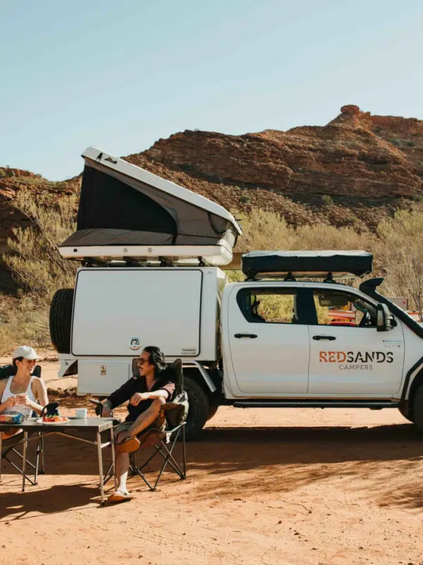 A man and woman sit on their camping chairs, sharing a coffee and snacks in front of their hired Redsands Camper vehicle. They are travelling across the Dampier Peninsula, towards Cygnet Bay Pearl Farm and have their tent pitched on the roof of the Redsands Camper vehicle. Surrounding them is the rocky terrain, red sand and native bush quintessential to the Kimberley region.