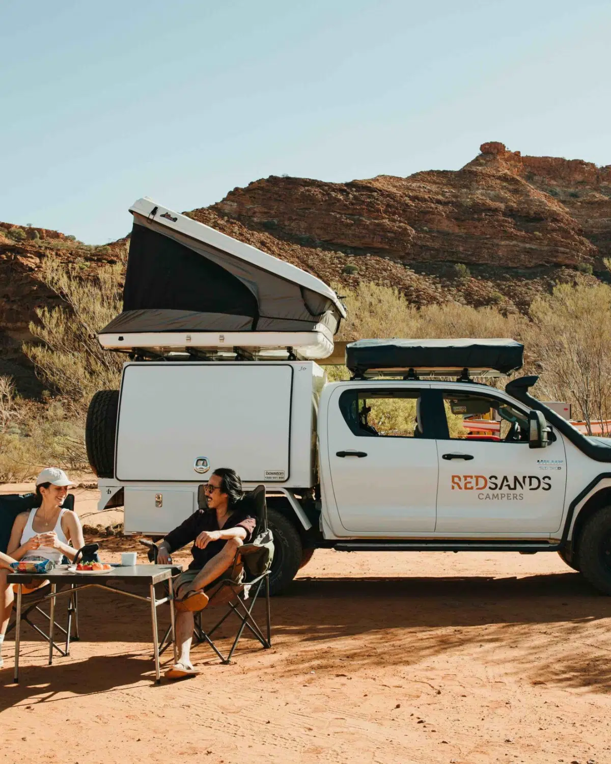 A man and woman sit on their camping chairs, sharing a coffee and snacks in front of their hired Redsands Camper vehicle. They are travelling across the Dampier Peninsula, towards Cygnet Bay Pearl Farm and have their tent pitched on the roof of the Redsands Camper vehicle. Surrounding them is the rocky terrain, red sand and native bush quintessential to the Kimberley region.
