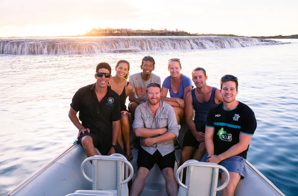 A group of guests smiling, sit on a boat after enjoying the Waterfall Reef Sea Safari. In the distance is the natural phenomenon that is Waterfall Reef and a stunning sunset.