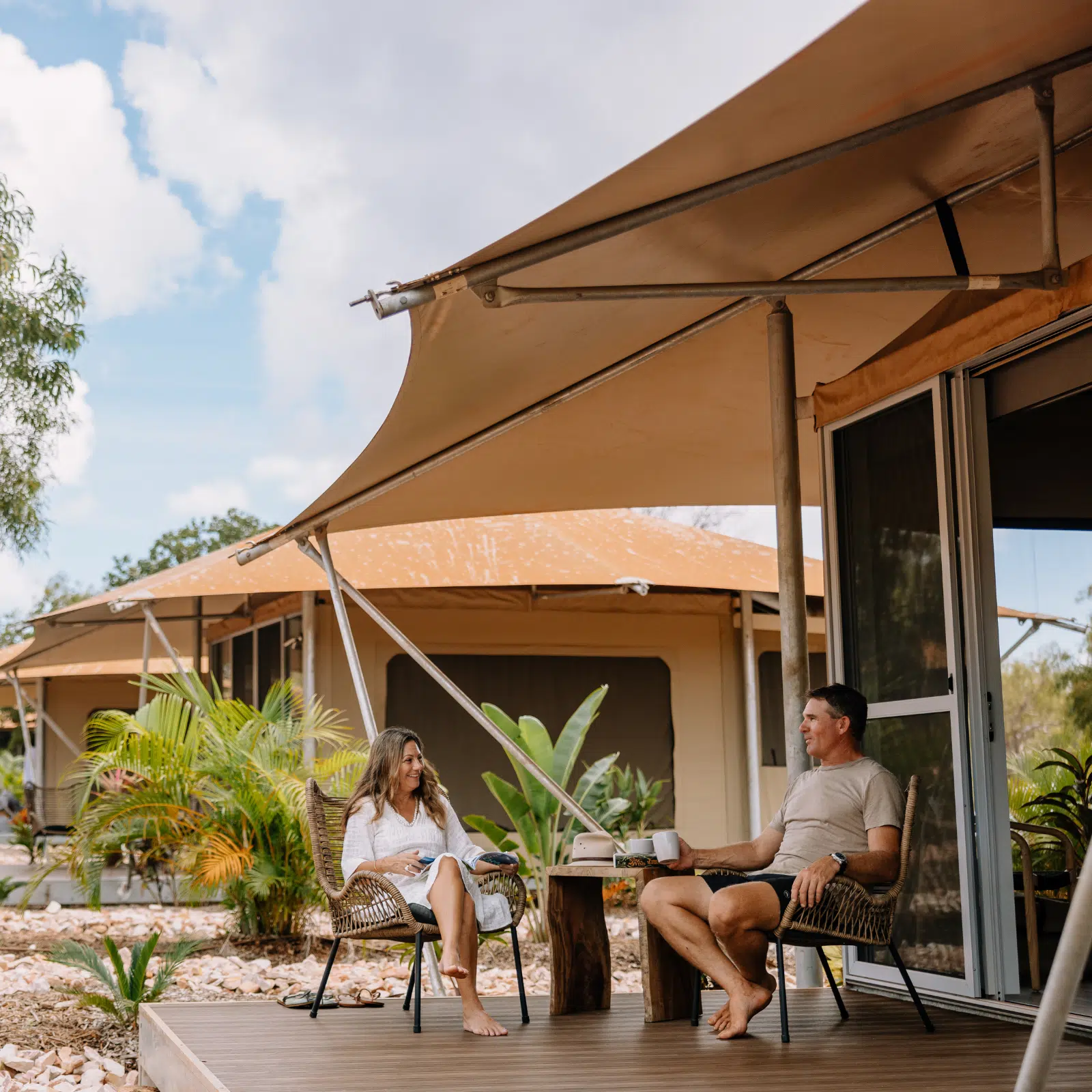 A couple sit on two rattan chairs, sharing a cup of coffee at their Pearler's Village Safari Tent accommodation. They are laughing, and enjoying the beautiful weather and serenity of Cygnet Bay Pearl Farm.