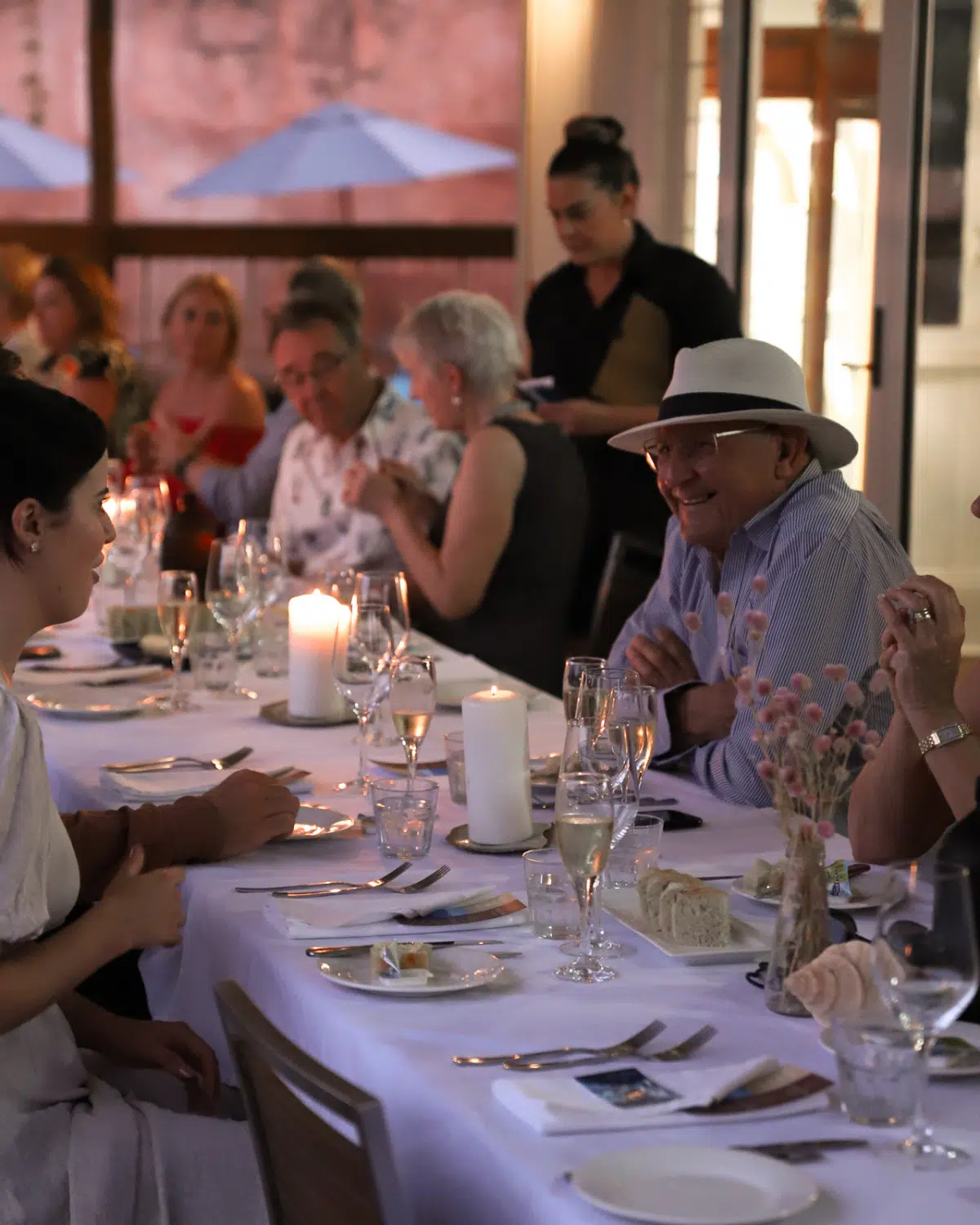 A group sit intimately at a table at sunset, overlooking the beautiful ocean of the Dampier Peninsula. The table is set with beautiful coastal elements of shells, and native florals, as well as candles to create a lovely, ambient setting.